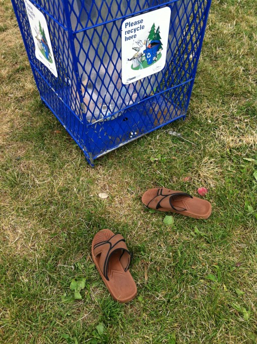 A pair of brown sandals on the grass near a recycle bin at the park.